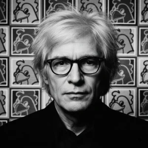 Retrato de Andy Warhol en blanco y negro, con fondo de patrón Pop Art multicolor Pollo Feliz.